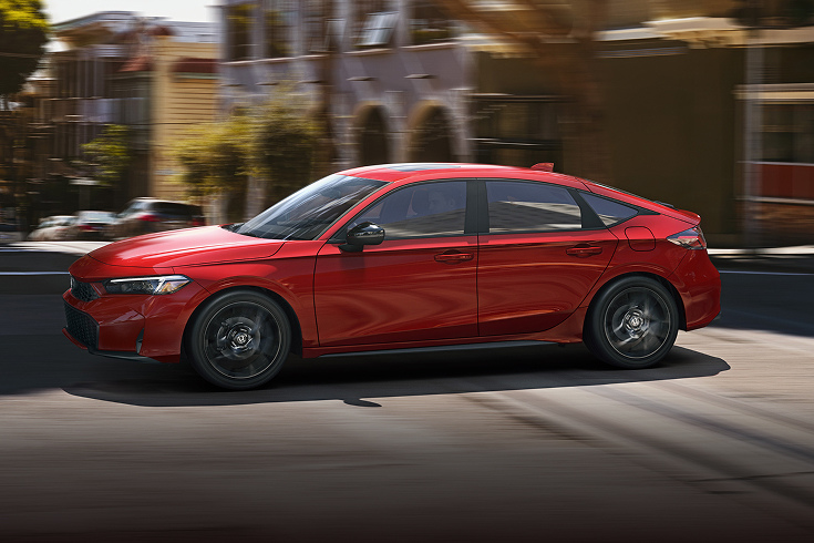 Perfil del Honda Civic Hatchback Sport Hybrid 2025 en Rallye Red conduciendo por una calle urbana (hatchback compacto).