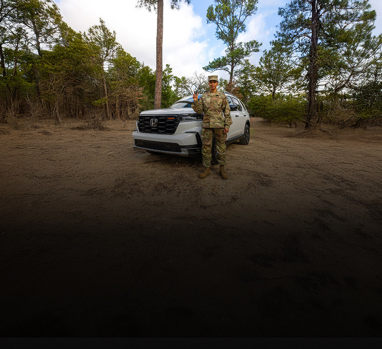 Parte delantera de la Honda Pilot TrailSport 2025 en Platinum White Pearl en un camino de tierra en el bosque con un soldado (SUV de tamaño completo).