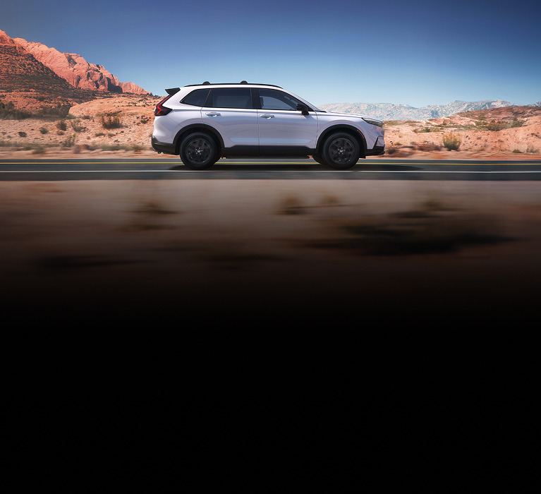 Perfil de la Honda CR-V Sport Touring Hybrid 2026 en Platinum White Pearl conduciendo por una carretera pintoresca (SUV crossover compacta).
